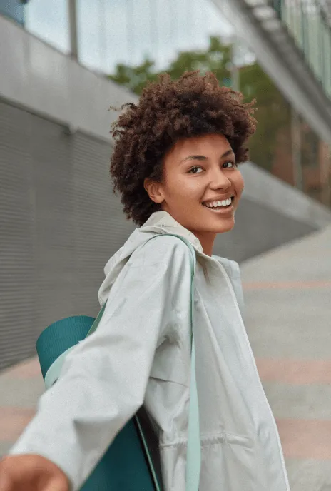 Mujer joven de cabello rizado sonriendo mientras carga su tapete de yoga.